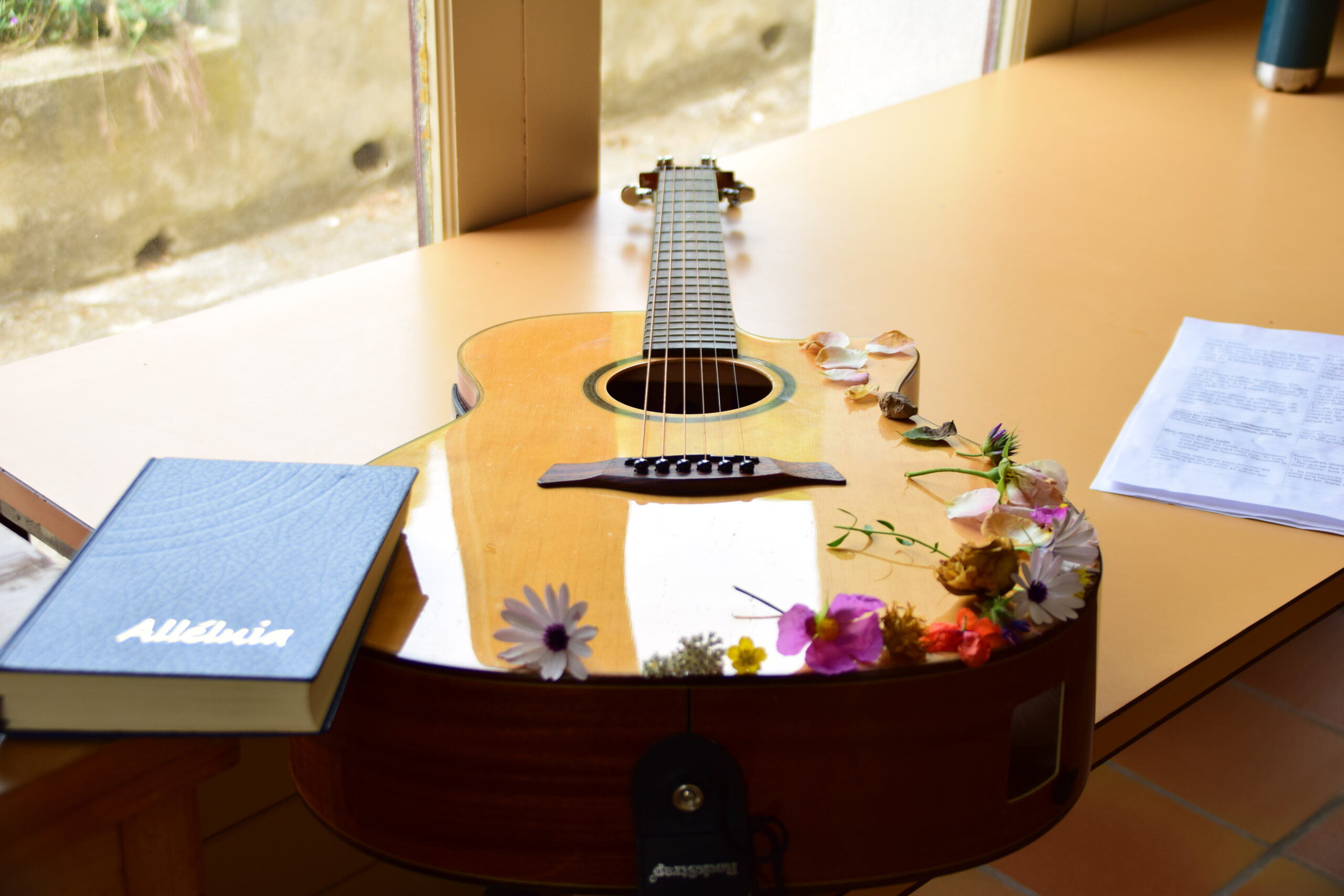 Une guitare est posée sur une table, il y a des fleurs dessus et un livre de chant